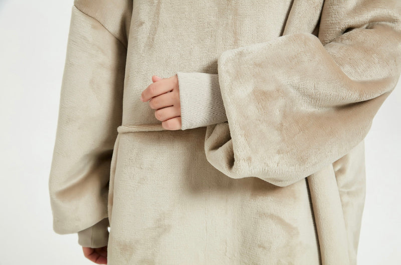 A close-up of a beige Cozify plush wearable blanket hoodie showing a person’s hand inside the front pocket, highlighting the soft and velvety fabric texture.