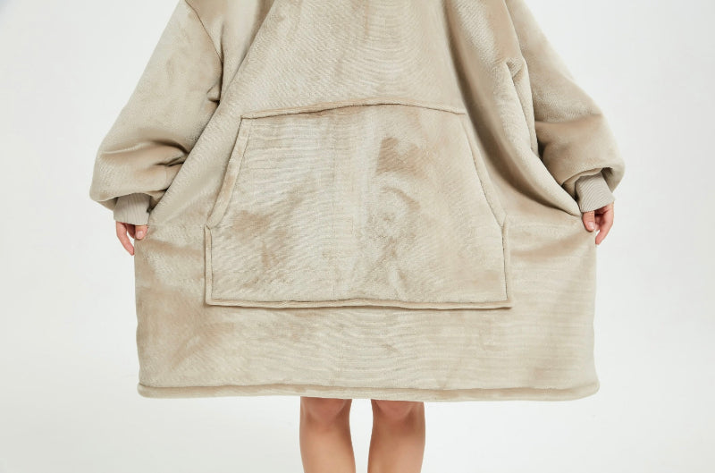 A close-up of a beige Cozify plush wearable blanket hoodie showing a person’s hand inside the front pocket, highlighting the soft and velvety fabric texture.