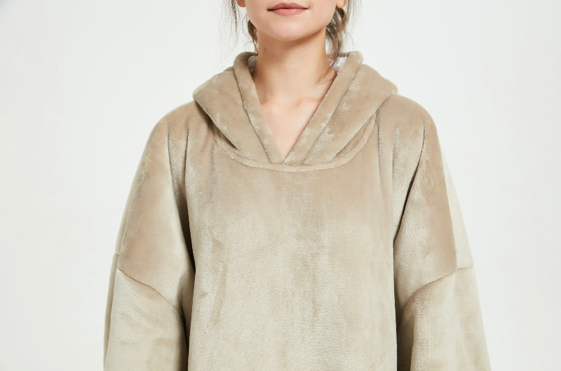 A close-up of a beige Cozify plush wearable blanket hoodie showing a person’s hand inside the front pocket, highlighting the soft and velvety fabric texture.