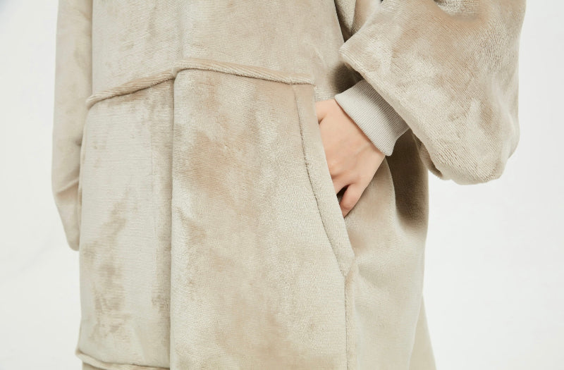 A close-up of a beige Cozify plush wearable blanket hoodie showing a person’s hand inside the front pocket, highlighting the soft and velvety fabric texture.