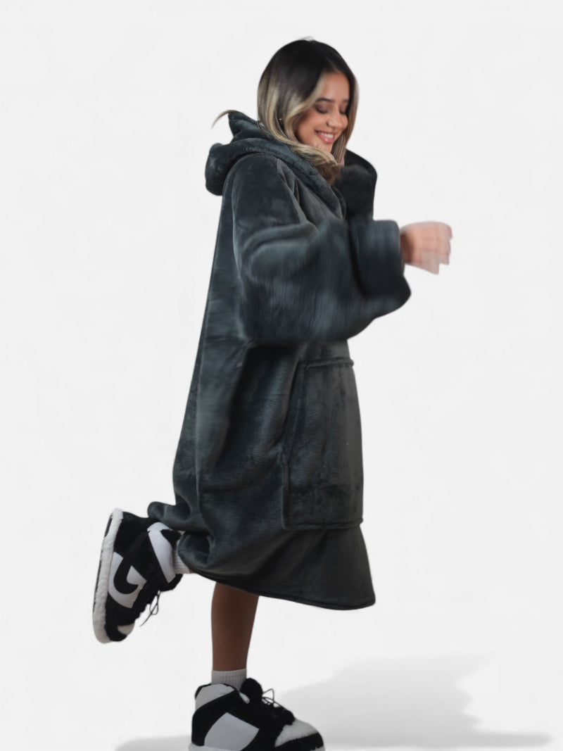 Side view of a smiling woman wearing a charcoal gray oversized Cozify sherpa-lined hoodie blanket with a front pocket, paired with black-and-white cozy slippers.