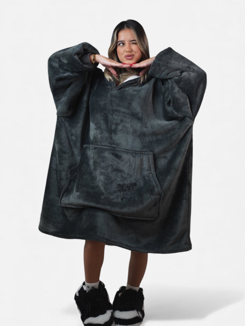 Woman playfully posing in a full-body shot wearing a charcoal gray oversized Cozify sherpa-lined blanket hoodie with front pocket and black-and-white fuzzy slippers.