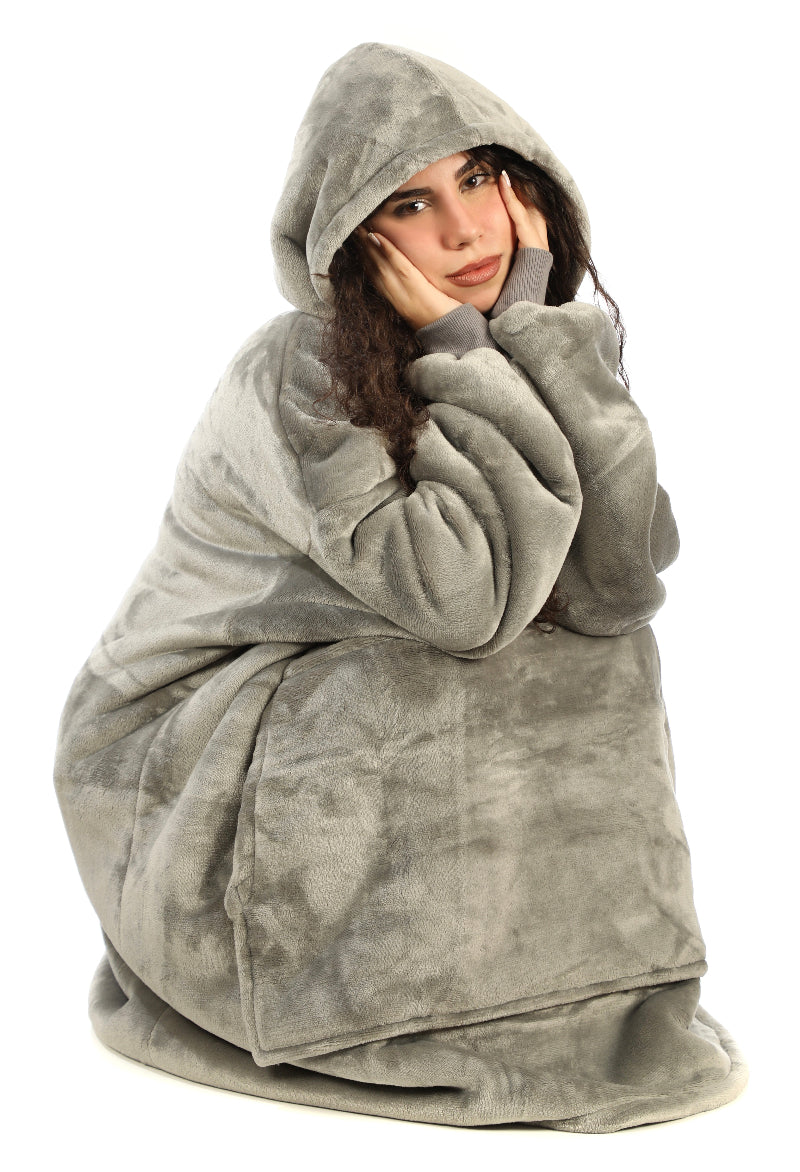 A woman sitting on the floor wearing a gray Cozify plush wearable blanket hoodie and matching gray slippers, facing forward and showing the soft, oversized design and cozy texture.