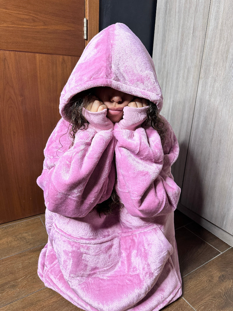 A woman wearing a pink Cozify plush wearable blanket hoodie sits on the floor with the hood up, resting her face in her hands and showing the soft, cozy fabric texture.