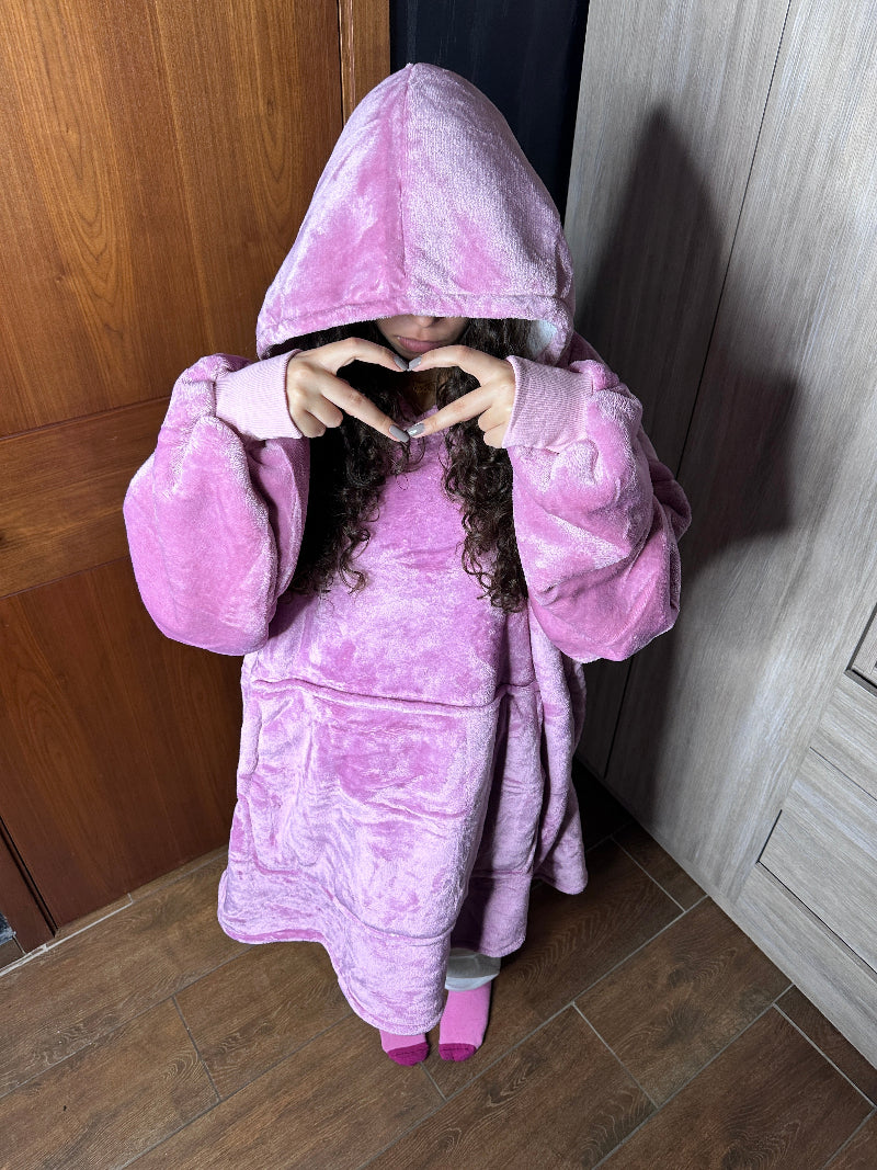 A top view of a woman wearing a pink Cozify plush wearable blanket hoodie, sitting on the floor with the hood up and her hands resting on her cheeks, showing the soft velvety texture.