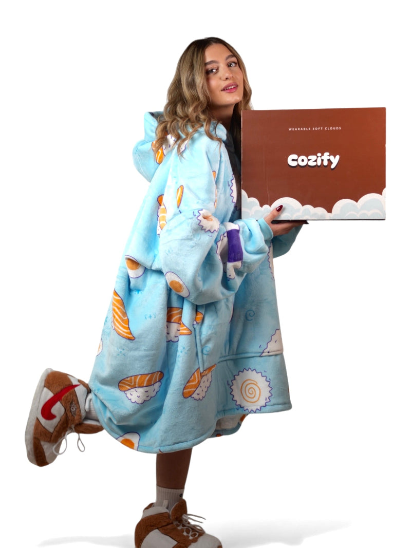 Smiling woman wearing a light blue sushi-print oversized wearable blanket hoodie while holding a brown Cozify box labeled “Wearable Soft Clouds,” posing on one leg against a white background.