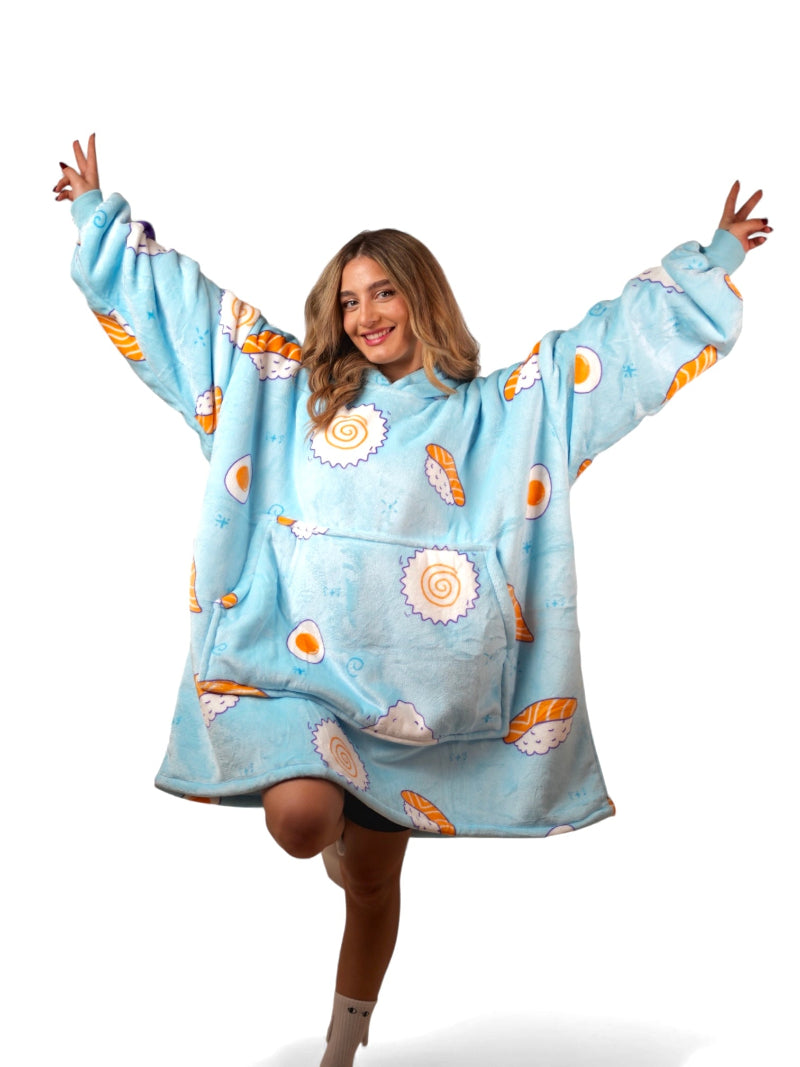 A smiling woman wearing a light blue oversized wearable blanket hoodie with a playful sushi print and a large front pocket, standing on one leg against a white background.